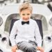 A boy sits on the grill of a luxury car dressed in formal wear with a bow tie | Household Staffing